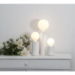 Drei weiße Leuchtenfuß-Mio-Lampen mit leuchtenden runden Glühbirnen stehen in unterschiedlichen Höhen auf einer weißen Kommode. Links davon bildet ein Blumenarrangement aus weißen Lilien und grünen Blättern einen schönen Kontrast zur schlichten weißen Wand.