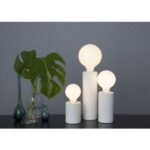 Drei moderne Leuchtenfuß - Mio Tischlampen in unterschiedlichen Höhen, jeweils mit einer runden weißen Glühbirne, stehen auf einer dunklen Fläche vor einer grauen Wand. Zwei grüne Glasvasen mit großen Monstera-Blättern komplettieren das minimalistische Dekor.