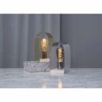 Zwei Tischlampen stehen auf einer Marmoroberfläche, jede mit einem gewölbten Metallrahmen – eine goldene und eine silberne –, in dem eine dimmbare LED-Kolbenbirne (3,2 W, 2100 K, Bernstein) steckt. Ein grauer Vorhang dient als Hintergrund.