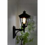 Die LED Kolben 3,6 W 2100 K Ø 6,4 cm Außenwandlaterne im Vintage-Stil ist mit einer leuchtenden Glühbirne auf einer weißen Verkleidung montiert, umgeben von einer weiß blühenden Pflanze darunter und einer verschwommenen grünen Landschaft im Hintergrund.