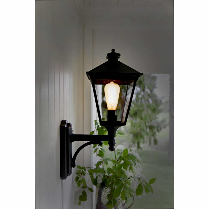 Die LED Kolben 3,6 W 2100 K Ø 6,4 cm Außenwandlaterne im Vintage-Stil ist mit einer leuchtenden Glühbirne auf einer weißen Verkleidung montiert, umgeben von einer weiß blühenden Pflanze darunter und einer verschwommenen grünen Landschaft im Hintergrund.