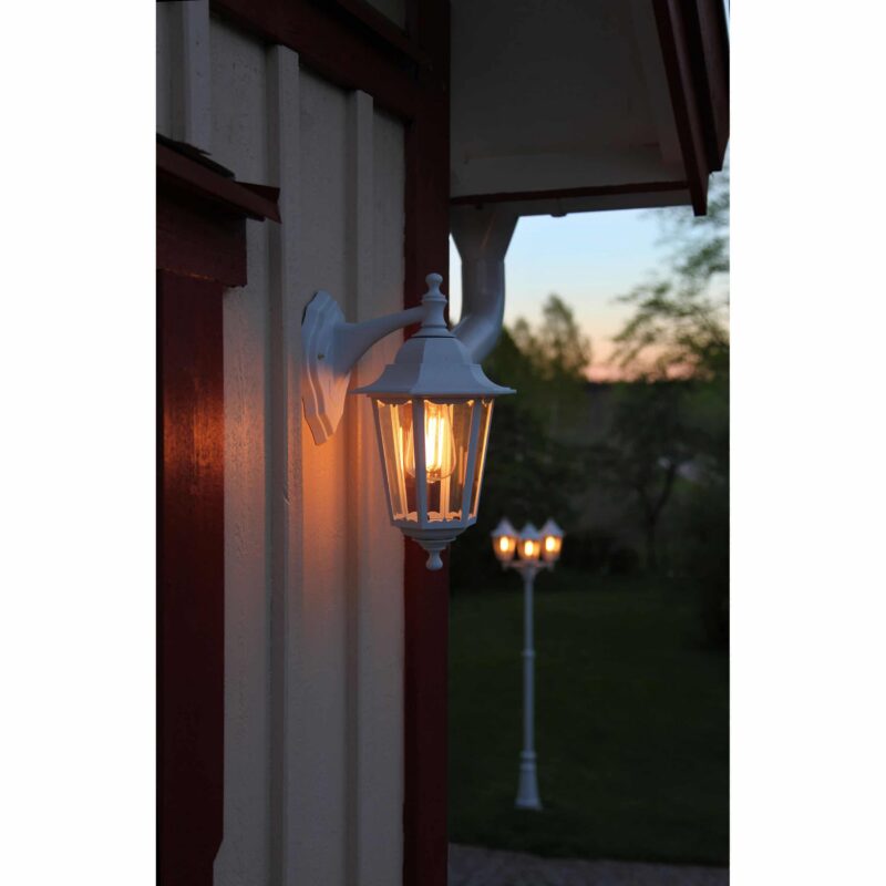 Eine LED Kolbenlaterne 3,6W 2100K Ø6,4cm beleuchtet in der Abenddämmerung die Außenseite eines Hauses. Im Hintergrund erhellt eine weitere ähnliche Laterne einen Garten und hebt die Silhouetten der Bäume vom Abendhimmel ab.