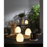 Ein gemütliches Esszimmer mit Nachtlicht-Solly-Lampen, die einen dunklen Tisch beleuchten. Schwarze Tassen stehen auf Untersetzern und auf einer Korbmatte liegen Holzstäbchen. Topfpflanzen und sanftes Licht verstärken die warme, einladende Atmosphäre.