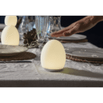 Eine Hand schwebt über der eiförmigen LED-Lampe Nachtlicht - Solly auf einem für eine Mahlzeit gedeckten Esstisch. Teller und Gläser sind ordentlich platziert, während Lichterketten die Umgebung schmücken. Die Lampe strahlt ein sanftes, warmes Licht aus und verstärkt die gemütliche Atmosphäre.