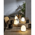 Ein Holztisch mit eiförmigen Nachtlichtern von Solly, vier schwarzen Tassen auf Untersetzern, einem Glas mit Grissini und einer Pflanze im Hintergrund. Gemütliches Ambiente mit sanfter Beleuchtung und einem Stuhl in neutralen Farben.