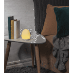 Eine gemütliche Schlafzimmerszene zeigt einen kleinen weißen Tisch neben dem Bett, geschmückt mit einem leuchtenden eiförmigen Nachtlicht – Solly. Das Ambiente wird durch zwei Bücher und ein Glas Wasser auf dem Tisch vervollständigt. Das Bett ist mit braunen Decken, einem grünen Kissen und einer grauen Strickdecke ausgestattet.