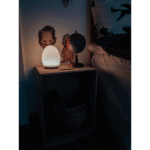 Eine gemütliche Nachttisch-Einrichtung umfasst ein leuchtendes, eiförmiges Nachtlicht – Solly, einen kleinen Globus und zwei illustrierte Tierausschnitte auf einem Holzregal. Darunter steht ein Weidenkorb. Das Bett ist mit Bettwäsche mit Katzenmuster ausgestattet, die eine warme, beleuchtete Atmosphäre schafft.