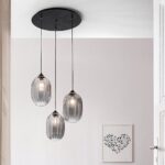 Die Pendelleuchte - Salva 3er Rondell präsentiert drei hängende durchscheinende Glaslampenschirme an schicken schwarzen Kabeln. Sie bildet einen Kontrast zu einer weißen Decke und einer beigen Wand und wird durch ein gerahmtes Kunstwerk mit einem Herzdesign aus schwarzen Formen ergänzt.