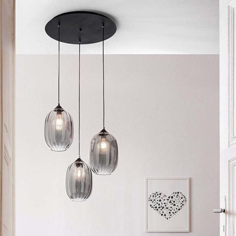 Die Pendelleuchte - Salva 3er Rondell präsentiert drei hängende durchscheinende Glaslampenschirme an schicken schwarzen Kabeln. Sie bildet einen Kontrast zu einer weißen Decke und einer beigen Wand und wird durch ein gerahmtes Kunstwerk mit einem Herzdesign aus schwarzen Formen ergänzt.