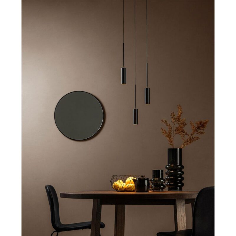 Ein moderner Essbereich verfügt über einen runden Spiegel an einer beigen Wand, mit drei Hängeleuchten - Tulpe Rondell über einem Tisch und schwarzen Stühlen. Auf dem Tisch stehen eine Vase mit Trockenblumen, eine dekorative Schale mit brennenden Kerzen und ein schwarzer Becher.