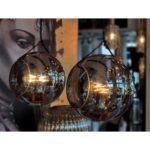 Zwei hängende, kugelförmige Glaslampen, die der eleganten Pendelleuchte Naru ähneln, verfügen über warm leuchtende Glühfäden. Sie sind vor einem unscharfen Hintergrund mit einem teilweise sichtbaren Graustufenporträt auf der linken Seite angebracht und erzeugen so eine gemütliche, künstlerische Atmosphäre.