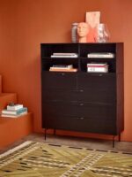 Vor einer orangefarbenen Wand steht eine schwarze Kommode mit offenen Regalen voller Bücher und Deko. Die Glass Bum-Vase steht neben einer Kopfskulptur und verleiht ihr Charme. Vorne liegt ein gemusterter Teppich, während links Stufen mit weiteren Büchern die gemütliche Szene vervollständigen.