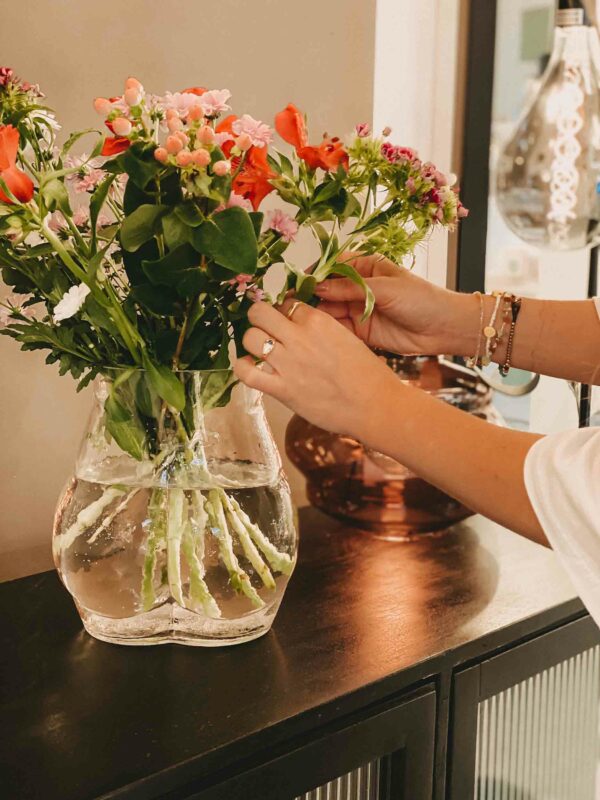 Eine Person arrangiert gekonnt einen lebhaften Strauß rosa, roter und orangefarbener Blumen in einer durchsichtigen, mit Wasser gefüllten Glasvase. Die elegante Vase steht auf einer dunklen Holzoberfläche mit einer sanft verschwommenen dekorativen Hängelampe im Hintergrund.