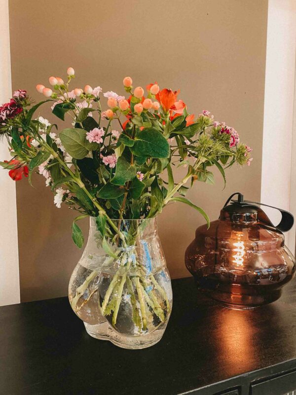 Eine Glass Bum-Vase mit rosa, orangefarbenen und roten Blumen und üppigen grünen Blättern steht elegant auf einem dunklen Tisch. Daneben steht eine braune transparente Glaslaterne mit Griff. Das Ganze steht vor einer zweifarbigen Wand, die den Charme der Ausstellung noch verstärkt.