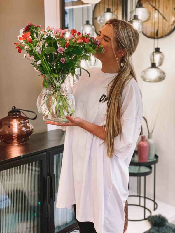 Eine Frau in einem übergroßen weißen Hemd hält einen großen, bunten Blumenstrauß in einer Vase – Glass Bum – im Innenbereich und steht vor einem Sideboard mit Dekoartikeln und Spiegeln. Deckenleuchten erhellen die Szene sanft und verleihen dem Raum Wärme.