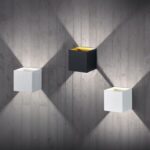 Drei Wandleuchten - Cube sind an einer strukturierten grauen Betonwand montiert, wobei zwei weiße Leuchten eine schwarze in der Mitte flankieren. Jede Leuchte strahlt ein weiches diagonales Licht aus, das dreieckige Muster bildet.