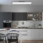 Die moderne Küche verfügt über eine geometrische Rückwand, weiße und dunkelgraue Schränke und einen eingebauten Ofen. Im Vordergrund steht ein weißer Tisch mit schwarzen Stühlen und einer Blumenvase. Offene Regale beherbergen Geschirr und Pflanzen, während helle schwarze Deckenleuchten von Frame (120 x 30 cm) den Raum erhellen.