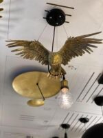 Die Pendelleuchte - Hedwig ist eine Deckenleuchte in Form eines Steinadlers mit ausgebreiteten Flügeln, der eine leuchtende Glühbirne in seinen Krallen hält. Sie verfügt über kreisförmige, goldfarbene Dekorationselemente an der Decke.