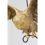 Die Pendelleuchte - Hedwig besteht aus einer goldenen Eulenskulptur mit detaillierter Federstruktur, die elegant auf einem schwarzen Metallhaken thront. Ihre Flügel sind weit ausgebreitet, wodurch ihr kompliziertes Design vor einem schlichten Hintergrund hervorgehoben wird.