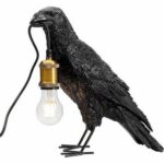 Die Tischleuchte - Black Crow besteht aus einer Rabenskulptur, die eine nicht angeschlossene Glühbirne mit einer goldenen Fassung im Schnabel hält. Der Vogel steht auf einer Oberfläche und lässt die Glühbirne an einer Schnur hängen, was ein einzigartiges Design ergibt.