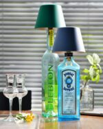 Zwei Schnapsflaschen mit Lampenschirmen veranschaulichen den Flaschenaufsatz Akku - Top 2.0 RGBW-Stil, darunter eine Leblon mit grünem Etikett und eine Bombay Sapphire mit blauem Etikett. In der Nähe schmücken zwei leere Gläser und eine kleine Blume den Holztisch neben einer Topfpflanze.