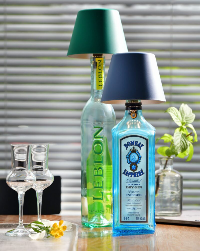 Zwei Schnapsflaschen mit Lampenschirmen veranschaulichen den Flaschenaufsatz Akku - Top 2.0 RGBW-Stil, darunter eine Leblon mit grünem Etikett und eine Bombay Sapphire mit blauem Etikett. In der Nähe schmücken zwei leere Gläser und eine kleine Blume den Holztisch neben einer Topfpflanze.
