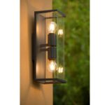 Die Außenwandleuchte - Tina ist eine wandmontierte Außenleuchte mit rechteckigem Metallrahmen und Glasplatten, in der zwei Glühbirnen untergebracht sind. Sie ist an einer orangefarbenen Wand mit verschwommenem Grün im Hintergrund befestigt.