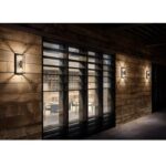 Eine moderne Holzfassade mit Glasfenstern gibt den Blick auf einen Essbereich frei. Drei Außenwandleuchten - Tina aus schwarzem Metall erhellen die Szene und werfen warmes Licht auf die Holzpaneele. Durch die Fenster sind Tische und Stühle sichtbar.