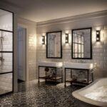 Ein stilvolles Badezimmer verfügt über zwei Waschbecken auf schwarzen Untergestellen, große Spiegel und Beleuchtung im Vintage-Stil von Außenwandleuchte - Tina. Der Boden ist mit gemusterten Fliesen ausgestattet, die Wände sind mit weißen U-Bahn-Fliesen verziert. Auf der rechten Seite befindet sich eine freistehende Badewanne und auf der linken Seite eine Glasdusche.