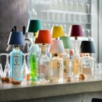 Flaschenaufsatz Akku - Top 2.0 RGBW-Lampenschirme zieren verschiedene Schnapsflaschen in einer Reihe auf der Bartheke, daneben liegen verstreut Korken und Gläser. Im Hintergrund sind Barutensilien zu sehen.