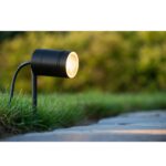 Der schwarze Gartenstrahler - Yona ist im Gras neben einem Steinweg installiert. Er erleuchtet mit einem warmen Licht und erzeugt einen verschwommenen grünen Hintergrund aus Büschen.