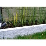 Hohe, dünne Bambusstäbe sind in ein Gartenbeet gepflanzt, das von Beton oder Stein begrenzt wird. Im Hintergrund befindet sich ein Holzzaun, und der Gartenstrahler - Yona-Strahler auf der linken Seite wertet die Szene auf. Im Vordergrund wächst üppiges grünes Gras, das zur ruhigen Atmosphäre beiträgt.