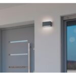 Ein modernes, minimalistisches Äußeres zeigt eine graue Tür mit horizontalen metallischen Akzenten. Darüber wirft die Wandleuchte - Up & Down Tegel einen subtilen Schein auf die weiße Wand. Rechts ist teilweise ein Fenster sichtbar.