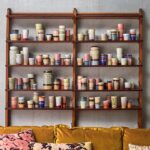 Auf Holzregalen sind eine Reihe farbenfroher Keramiktassen ausgestellt, darunter der bezaubernde Kaffeebecher „Vintage Nature“ von HK Living. Die Designs sind gestreift, gesprenkelt und in Pastelltönen gehalten. Unter den Regalen steht ein senfgelbes Sofa mit geblümten Kissen vor einer schlichten Wand.