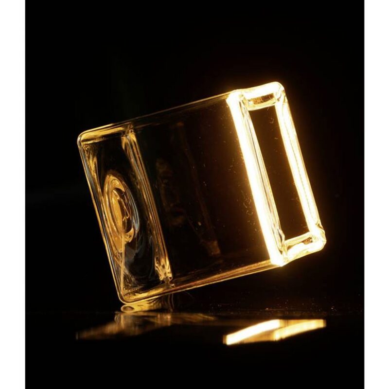 Ein transparenter Floatlight Cube 6W dimmbar klar 12,5cm steht auf seiner Kante und wird von einer warmen, goldenen Leuchtmitte vor einem dunklen Hintergrund angestrahlt, wodurch ein leuchtender Effekt entsteht.