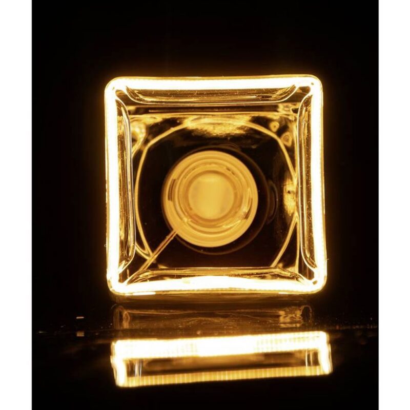 Eine Nahaufnahme der leuchtenden, quadratischen Leuchtmitte Floatlight Cube 6W dimmbar klar 12,5cm vor einem schwarzen Hintergrund, die mit ihrem warmen Schein einen hellen gelben Farbton und ein symmetrisches geometrisches Muster erzeugt.