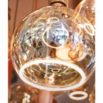 Nahaufnahme einer kugelförmigen Pendelleuchte aus Glas mit einem kreisförmigen Leuchtmittel Floatlight Globe 5W dimmbar klar Ø20cm im Inneren. Die reflektierende Oberfläche verzerrt die Umgebung mit verschiedenen Formen und Lichtern, während im Hintergrund ähnliche Leuchtmittel sichtbar sind.