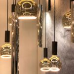 Hängende Pendelleuchten mit runden Glasabdeckungen und das Leuchtmittel Floatlight Globe 4W dimmbar gold Ø15cm strahlen ein warmes Licht vor einer strukturierten grauen Wand aus. Mit schwarzen Akzenten sind sie in unterschiedlichen Höhen angeordnet und schaffen ein elegantes und modernes Ambiente.