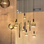 Ein Satz dimmbarer Floatlight Globe 4W-Pendelleuchten in Gold mit freiliegendem Leuchtmittel, in verschiedenen metallischen Ausführungen. Sie hängen in unterschiedlichen Höhen vor einer strukturierten beigen Wand und sorgen für eine elegante und moderne Atmosphäre.