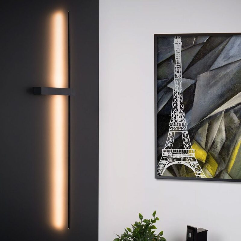 Eine schlanke Wandleuchte - Slimline schmückt eine schwarze Tafel neben einem geometrischen Eiffelturm-Gemälde an einer makellos weißen Wand. Darunter verleiht eine kleine Pflanze dem modernen Dekor einen Hauch von Grün.