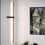 An einer Wand hängt eine Wandleuchte - Slimline mit einem gerahmten abstrakten Uhrturm-Gemälde, das an Big Ben erinnert. Das Kunstwerk mit geometrischen Formen in Schwarz, Weiß und Gelb wird durch eine kleine Pflanze am Fuß des Rahmens hervorgehoben.