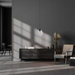Ein minimalistisches Interieur besteht aus einem dunklen Sideboard, einem beigen Sessel und einer eleganten Pendelleuchte - Bubble 1er (M). Sonnenlicht erzeugt geometrische Muster an den dunklen Wänden. Zur Dekoration gehören Bücher, Vasen und Pampasgras. Im Hintergrund ist ein Essbereich zu sehen.