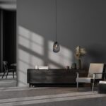 Ein modernes Interieur zeigt ein dunkles Sideboard vor einer grauen Wand, darüber hängt die Pendelleuchte - Bubble 1er (L) mit dekorativen Vasen und Gräsern. Neben dem Sideboard steht ein hell gepolsterter Sessel, während Sonnenlicht durch ein Fenster Schatten wirft; im Hintergrund stehen ein Tisch und ein Stuhl.