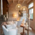 Ein gemütliches Wohnzimmer verfügt über einen weißen Stuhl, hölzerne Beistelltische mit dekorativen Vasen und drei Pendelleuchten „Pendelleuchte - Fawn“, die Kerzen und eine Lampe zum Leuchten bringen. Über dem Stuhl hängt eine Decke, während Sonnenlicht durch ein Fenster fällt und Wärme verbreitet.