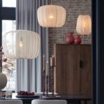 Ein Esszimmer mit drei beigefarbenen Noemi-Hängelampen, einem mit Kerzen geschmückten Holztisch und roten Dekovasen auf einem Schrank. Der Raum verfügt über eine Ziegelwand und ein großes Fenster mit hellgrauen Vorhängen.