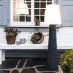 Eine gemütliche Außenszene zeigt die 160 cm große Stehleuchte Ronda neben einer weißen Bank mit geflochtenen Blumenkörben. Im Hintergrund ist ein Fenster mit Fensterläden und im Vordergrund eine kleine Topfpflanze zu sehen.