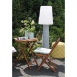 Eine Außeneinrichtung umfasst einen Holztisch und einen Stuhl mit weißen Kissen neben einer hohen 160 cm Ronda Stehleuchte. Ein gelb gemusterter Pouf steht auf dem dunklen Holzdeck, umgeben von grünem Laub und weißen Blumen, die eine ruhige Kulisse bilden.