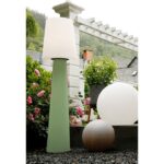 Eine große, grüne Ronda-Stehleuchte (160 cm) mit weißem Schirm steht zwischen rosa Rosen und Grünpflanzen. Daneben stehen eine weiße Kugellampe und eine kleinere braune Kugel, alles vor dem Hintergrund einer weißen Pergola und einer Steinmauer mit einem Kamin.