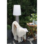 Eine gemütliche Außenumgebung umfasst einen Holzstuhl mit einer dicken weißen Strickdecke. Auf dem Tisch stehen eine Zeitschrift und eine blaue Kaffeetasse. Die 160 cm große Ronda Stehleuchte steht elegant neben dem Stuhl, umgeben von üppigem grünem Laub im Hintergrund.