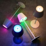 Vier Akku LED Tischleuchten mit RGB-Farben – Rosa, Grün, Blau und Weiß – sind auf einer dunklen Oberfläche angeordnet. Jede Lampe hat einen einfachen konischen Schirm und einen runden Sockel und bietet ein modernes und farbenfrohes Display.